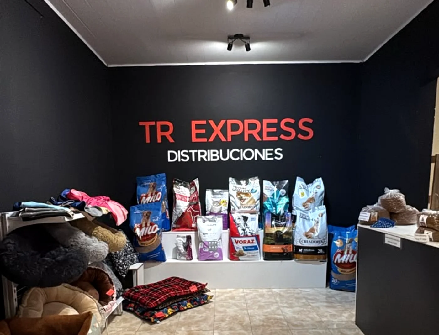 Interior del local de TR Express: pared negra con el cartel TR Express Distribuciones, estanterías con bolsas de alimento y camas para mascotas.